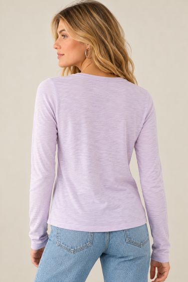 A woman wearing a long sleeve crew neck tee in lavender with blue jeans on a neutral background.
