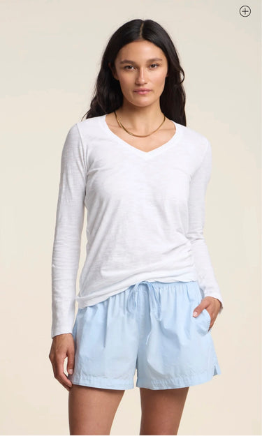 A woman modeling a white long-sleeve V-neck top and light blue drawstring shorts against a plain beige studio background.