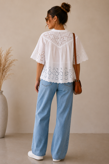 Woman wearing a white lace top and blue jeans against a beige wall.
