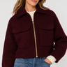 A woman wearing a cropped, deep burgundy bouclé shearling jacket with a gold zipper, paired with blue jeans and a cream ribbed sweater, against a light gray studio background.