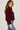 A woman wearing a cropped, long-sleeve sherpa jacket in deep burgundy with gold-tone zipper and button details, paired with blue jeans, photographed against a neutral light gray studio background.