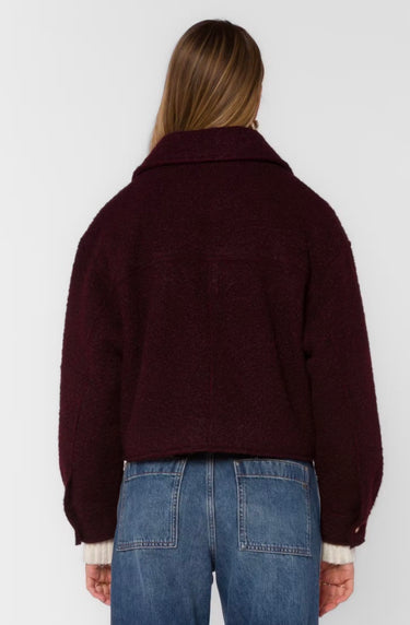 A woman viewed from the back wearing a cropped, textured deep burgundy jacket and blue jeans against a plain light gray studio background.