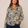A woman modeling a long-sleeve button-up blouse with an abstract leopard-like print in beige, black, and olive green, paired with dark blue jeans against a plain beige studio background.