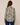 A woman viewed from the back wearing a long-sleeve button-up shirt with a beige, black, and brown floral pattern, paired with blue jeans, against a solid beige background.