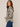 A woman wearing a beige button-up blouse with a navy blue floral pattern, paired with blue jeans, photographed against a solid light beige studio background.