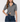 A woman modeling a short-sleeve gray cable-knit cropped cardigan with a contrasting dark blue collar and gold buttons, paired with dark blue jeans against a light gray studio background.