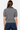 A woman viewed from the back wearing a gray short-sleeve cable-knit polo-style top with a blue collar, paired with dark blue jeans, against a plain light gray studio background.