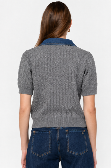 A woman viewed from the back wearing a gray short-sleeve cable-knit polo-style top with a blue collar, paired with dark blue jeans, against a plain light gray studio background.