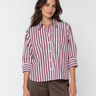 A woman modeling a long-sleeve button-up shirt with vertical burgundy and white stripes, paired with brown trousers, against a light gray studio background.