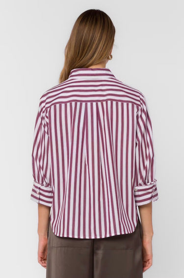 A woman viewed from the back wearing a long-sleeve button-up shirt with vertical burgundy and white stripes, paired with brown trousers, against a light gray studio background.