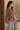 A woman wearing a brown long-sleeve blouse with a beige and white floral pattern, paired with light blue jeans, standing in a warm-toned indoor setting next to a wooden door and a potted plant.