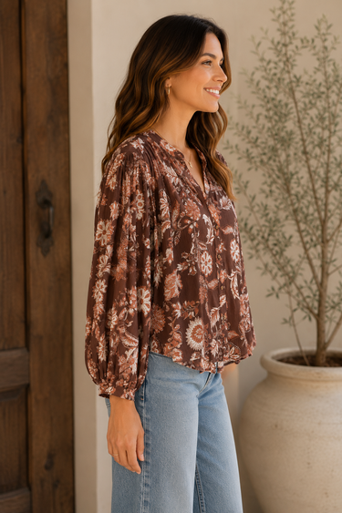 A woman wearing a brown long-sleeve blouse with a beige and white floral pattern, paired with light blue jeans, standing in a warm-toned indoor setting next to a wooden door and a potted plant.