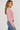 A woman wearing a fuzzy, long-sleeve cropped sweater in light pink, paired with blue jeans, photographed against a plain light gray studio background.