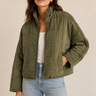 A woman wearing an olive green quilted jacket over a white ribbed top and light blue jeans, standing against a plain beige studio background.