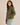 A woman wearing an olive green quilted jacket and light blue jeans, photographed from the side against a plain beige studio background.