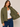 A woman wearing an olive green quilted jacket over a white ribbed tank top and blue jeans, holding the jacket open to show the interior lining against a plain beige studio background.