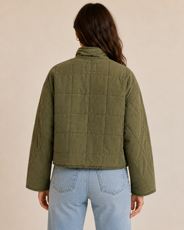 A woman viewed from the back wearing an olive green quilted jacket and light blue jeans, set against a plain beige studio background.