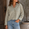A woman wearing a long-sleeve button-up shirt with vertical olive green and cream stripes, paired with light-wash blue jeans, standing in front of a rustic wooden door and a potted plant.