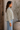 A woman wearing a beige and white vertically striped button-down shirt with light blue jeans, standing in profile next to a rustic wooden door and a potted plant.