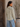 A woman wearing a cream and olive green vertically striped long-sleeve blouse with a band collar, paired with light blue jeans, standing in front of a rustic wooden door and a potted plant.