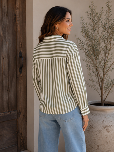 A woman wearing a cream and olive green vertically striped long-sleeve blouse with a band collar, paired with light blue jeans, standing in front of a rustic wooden door and a potted plant.