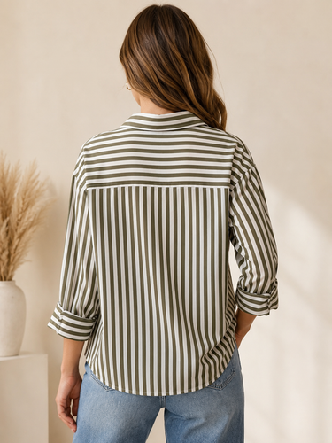 A woman viewed from behind wearing a striped button-up shirt with vertical stripes in olive green and off-white, paired with light blue jeans, set against a warm beige background with dried grass decor.