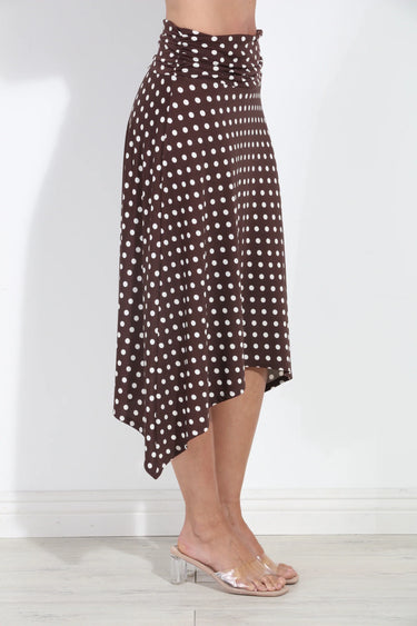 A woman wearing a brown and white polka dot high-waisted wrap skirt with an asymmetrical hem, paired with clear heeled mule sandals, standing against a white wall.