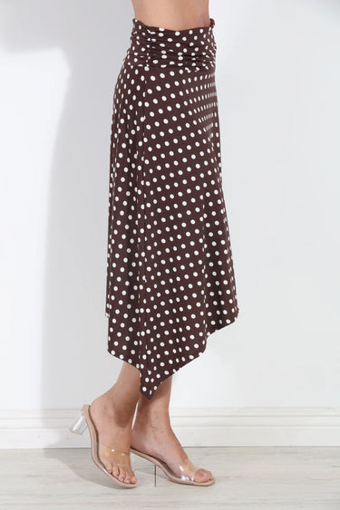 A brown midi skirt with a white polka dot pattern, shown on a model wearing clear high-heeled sandals against a white wall.