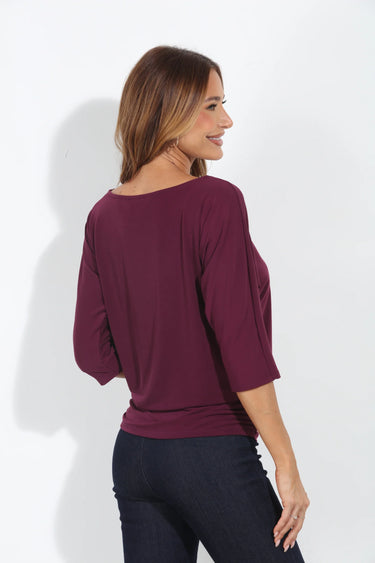 Plum off-the-shoulder three-quarter sleeve top with a relaxed draped fit, styled with dark wash jeans on a model.