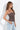 A woman wearing a brown spaghetti-strap camisole top with white polka dots, paired with light-wash blue jeans, standing against a white studio background.