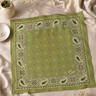 An olive green bandana with a white paisley border and small white floral motifs, styled flat on a beige fabric background with dried flowers and a small dish.