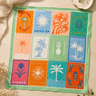 A square patchwork-style cotton bandana laid flat on a beige surface, featuring a grid of colorful tropical and sun motifs in orange, blue, pink, green, and teal. The cloth is styled with draped cream fabric, a small dish, and dried flowers around it.