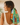A woman shown from the back wearing a colorful patchwork-patterned hair scarf tied in her ponytail, paired with a white sleeveless top.