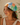 A woman wearing a colorful patchwork-print headscarf with tropical and beach-themed designs, styled with a gold pendant necklace and earrings in a warm, neutral indoor setting.
