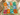 A square patch bandana with a colorful patchwork design featuring palm trees, suns, waves, and tropical motifs, laid out on a beige stone surface with decorative props.
