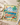 A square beach-themed scarf laid flat on a light beige fabric surface. The scarf features a colorful tropical illustration with palm trees, a blue ocean, mountains, and the words 'Paradise BEACH' in red and gold lettering.