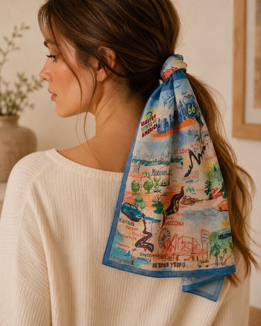 A woman wearing a cream ribbed knit sweater, shown from the back in profile. Her brown hair is tied back in a ponytail with a square cotton scarf featuring a colorful vintage map illustration of Route 66 and Southwest US landmarks.