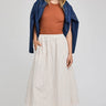 Woman wearing a rust top, white pleated skirt, and blue jacket on a plain background