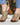 A woman wearing open-toe block-heel sandals with metallic champagne-gold textured uppers, bronze-tone metal studs, and brown wooden soles