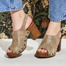 A woman wearing open-toe block-heel sandals with metallic champagne-gold textured uppers, bronze-tone metal studs, and brown wooden soles