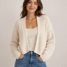 A woman wearing a cream-colored, open-front chunky knit cardigan over a matching cream tank top and blue jeans, standing against a plain light beige studio background.