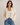 A woman wearing a cream-colored, open-front chunky knit cardigan over a matching cream tank top and blue jeans, standing against a plain light beige studio background.