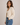 A woman wearing a cream open-knit cardigan over a beige top and blue jeans, standing against a plain light beige studio background.