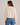 A woman shown from the back wearing a cream-colored, chunky knit cropped sweater and blue jeans against a plain light gray studio background.