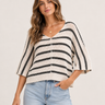 A woman wearing a cream and black horizontally striped knit sweater with elbow-length sleeves and a V-neck, paired with light blue jeans, against a plain light beige background.
