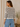A woman shown from the back wearing a cream knit sweater with black horizontal stripes and light blue jeans, standing indoors next to a neutral wall and a potted plant.
