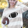 A smiling blonde woman outdoors wearing a cream and beige horizontally striped knit sweater with a large black football graphic on the chest, holding a brown football.