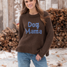 Woman wearing a brown knit “Dog Mama” sweater with light blue lettering and relaxed blue jeans in a rustic outdoor setting.