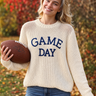 A woman holding a football wearing a cream-colored, chunky-knit long-sleeve sweater  with “GAME DAY” embroidered on the front and blue jeans, photographed from behind in an outdoor autumn park with colorful fall foliage.