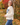 A woman wearing a cream-colored, chunky-knit long-sleeve sweater  with “GAME DAY” embroidered on the front and blue jeans, photographed from behind in an outdoor autumn park with colorful fall foliage.